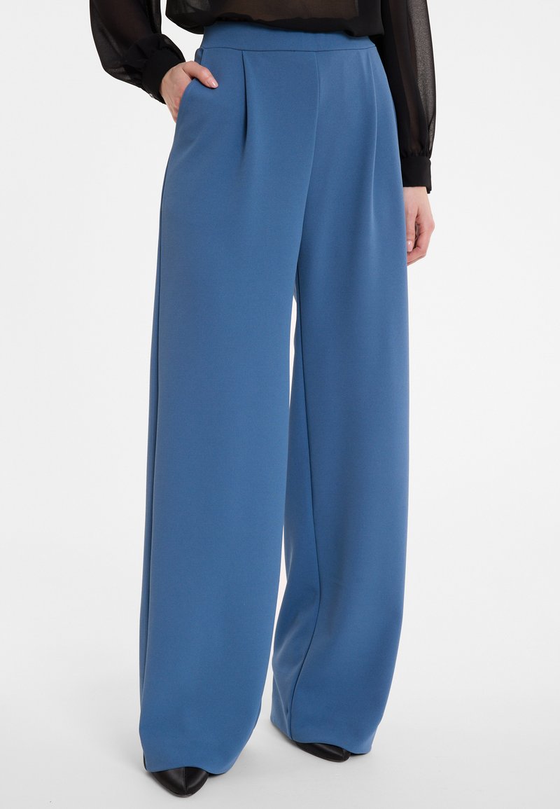 Blauwe wijde broek met een gladde, gestructureerde stof. Heeft plooien, zijzakken en een hoge taille. Gestyled met zwarte schoenen.