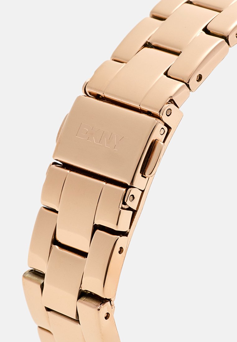 Pulsera de reloj de metal dorado con segmentos enlazados, que cuenta con un cierre y el logotipo grabado de DKNY, textura suave y acabado pulido.
