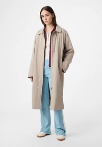 Lysebeige lang frakke med krave, med front lynlås og to sidelommer. Kombineret med lyseblå bukser og hvide sko.