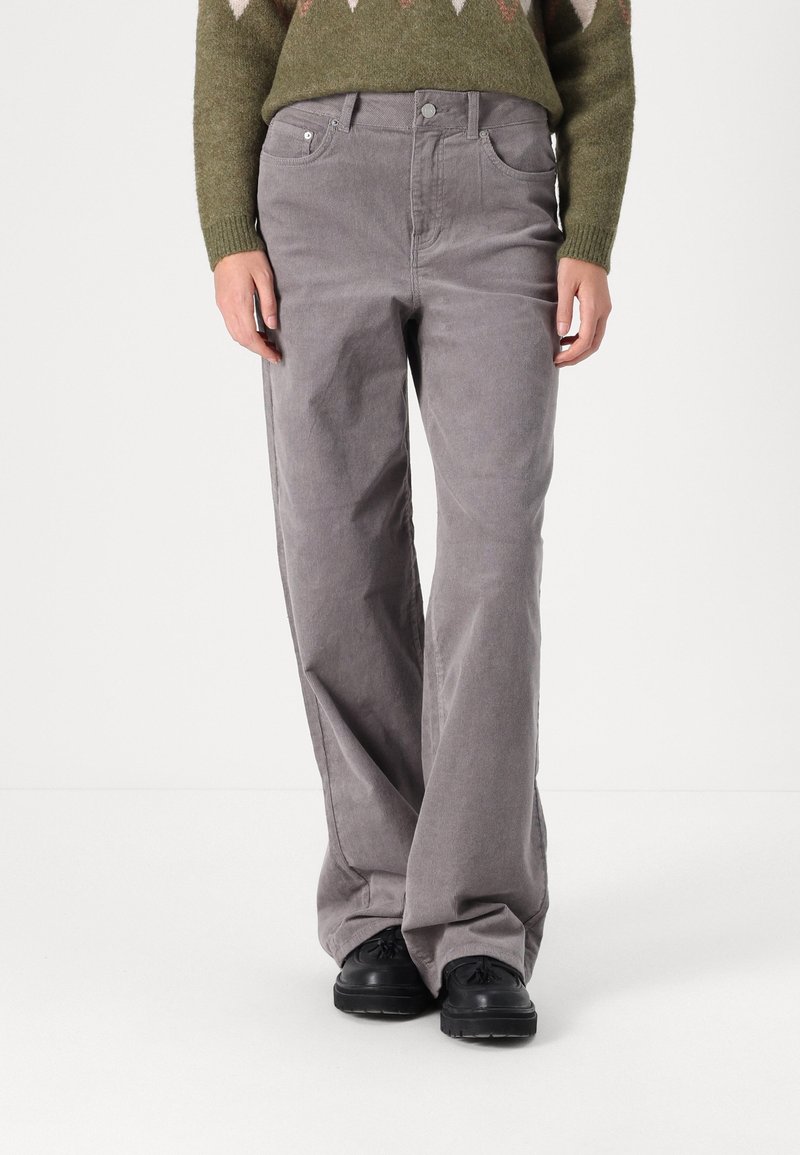 Personne portant un pantalon ample gris, des chaussures noires et un pull vert olive, debout sur un fond clair et uni.
