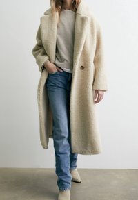 Manteau en laine texturée couleur crème, à coupe ample, larges revers et fermeture par boutons, porté sur un jean bleu clair et des bottines beige.