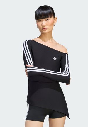 Donna con top nero Adidas scollato sulle spalle con strisce bianche sulle maniche e pantaloncini neri abbinati, in piedi con le braccia incrociate.