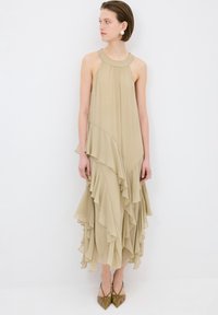 Beiges, fließendes Kleid mit Halterneck, versehen mit schichtweisen Rüschen am Saum und transparenter Stoffstruktur. Kombiniert mit spitzen, hellbraunen Schuhen.