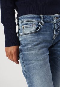 Verblasste blaue Denim-Jeans mit einem klassischen Fünf-Taschen-Design. Sie verfügen über einen Reißverschluss und dezente Nähte, kombiniert mit einem dunkel gestrickten Pullover.