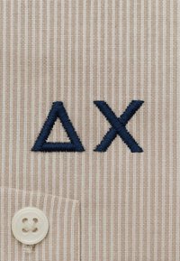Tessuto beige a righe con un bottone bianco e simboli ricamati blu navy: un triangolo (Δ) e una "X", posizionati sopra l'area della tasca.