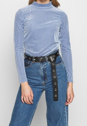 Frau trägt ein hellblaues Samtstreifen-Langarmshirt, das in eine hoch taillierte blaue Jeans mit schwarzem Ösengürtel gesteckt ist.