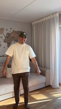 White short-sleeve t-shirt with a pocket, brown pants, standing on a wooden floor. Background features a light-colored sofa and curtains.