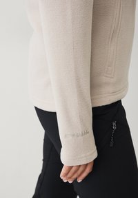 Giacca in pile beige chiaro con superficie texturizzata, dotata di una tasca con zip e logo ricamato sul polsino. Abbinata a pantaloni neri.