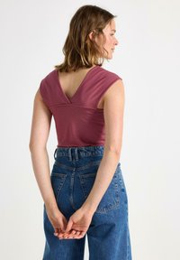 Bezramenný top v lila látce s designem V-výstřihu na zadní straně, v kombinaci s džínovými kalhotami s vysokým pasem v modré barvě, které mají zadní kapsy.