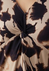 Blusa de manga corta en tejido suave con una base beige y un patrón de tie-dye marrón oscuro. Presenta un frente envolvente con un detalle de nudo en el centro.
