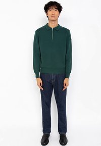 Polo a maniche lunghe verde scuro con colletto con zip, trama a coste e design semplice. Abbinato a jeans di denim blu e scarpe nere.