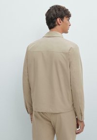Jonge man gezien van achteren, gekleed in een beige lange mouwen shirtjack en bijpassende broek tegen een eenvoudige lichte achtergrond.