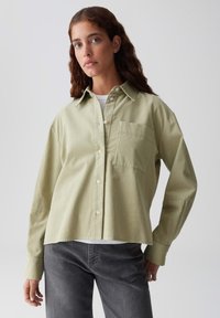 Frau mit welligem braunem Haar, die ein hellgrünes Hemd über einem weißen Top und einer dunkelgrauen Jeans trägt, steht vor einem einfarbigen, hellen Hintergrund.