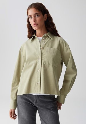 Woman with wavy brown hair wearing a light green button-up shirt over a white top and dark gray jeans, standing against a plain light background.