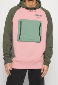 Person klädd i en rosa och olivgrön Nike hoodie med en stor grön dragkedjefick i fram och svarta byxor, stående mot en enfärgad bakgrund.