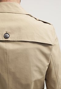 Trench coat beige con uno spacco posteriore, dettagli con bottoni neri e texture liscia, con cuciture visibili sulle cuciture.