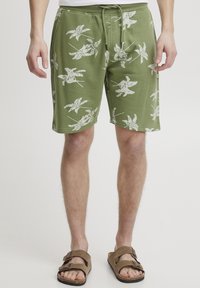 Pantaloni corti verdi con coulisse e motivi a foglia bianchi, realizzati in tessuto morbido, presentano tasche laterali e una vestibilità rilassata, abbinati a sandali marroni.