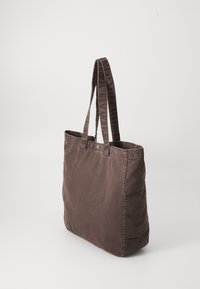 Brown canvas tote bag with dual fabric handles, rectangular shape, and visible stitching. Features a gold dot accent near the top.