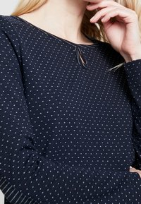 Granatowy longsleeve z małymi białymi trójkątnymi wzorami, okrągłym dekoltem z detalem w kształcie łezki i ściągaczami przy mankietach.