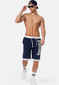 Navy blue swim shorts with white accent, elastic waistband, and drawstring. Features a large brand logo on the side. White trim at the hem.