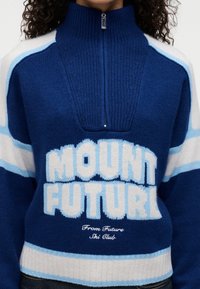 Πλεκτό πουλόβερ με ψηλό γιακά, κυρίως ναυτικό μπλε με λευκές και ανοιχτό μπλε ρίγες. Φέρει μεγάλο κείμενο "MOUNT FUTURE" και κεντημένο λογότυπο.