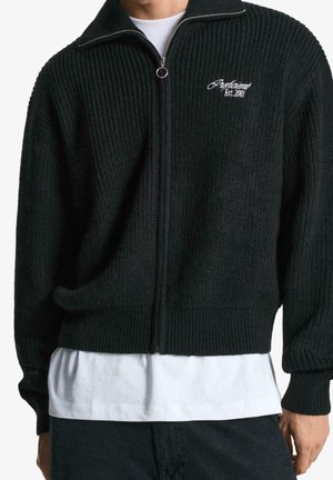 Mężczyzna ubrany w czarny, prążkowany sweter na zamek z haftem "Proficient Est. 2001" na piersi, noszony na białej koszulce.