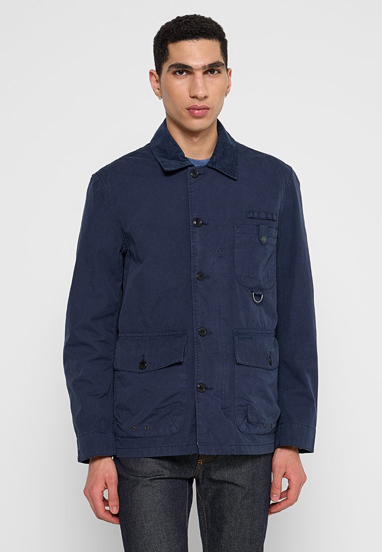 Giacca leggera blu navy con colletto in velluto a coste, multiple tasche, bottoni frontali e un dettaglio a anello sul petto.