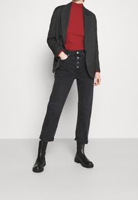 Person, der ein schwarzes gestreiftes Blazerjackett, einen roten Rollkragenpullover, hochtaillierte schwarze Jeans mit Knöpfen und schwarze Knöchelstiefel trägt und mit einer Hand in der Tasche steht.