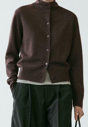 Cardigan marron foncé déboutonné sur une chemise grise, associé à un pantalon noir ample, main dans la poche, tenant une sangle noire.