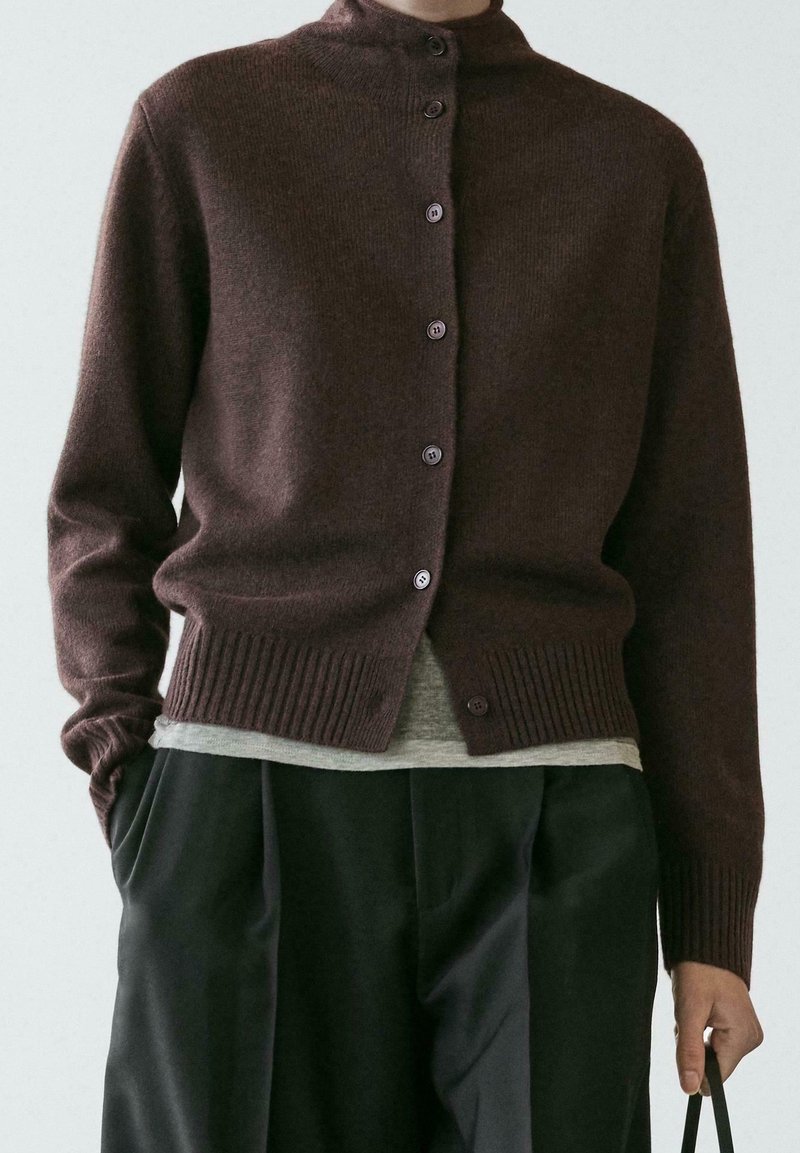Cardigan marron foncé déboutonné sur une chemise grise, associé à un pantalon noir ample, main dans la poche, tenant une sangle noire.