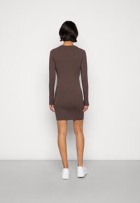 Brązowa, dopasowana sukienka z długim rękawem, do kolan, o prążkowanej fakturze, z okrągłym dekoltem, zestawiona z białymi sneakersami, widok od tyłu.