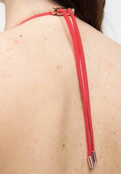 Κόκκινο τοπ halter δεμένο πίσω σε έναν ώμο με φακίδες, με χρυσές μεταλλικές άκρες στα λουράκια.