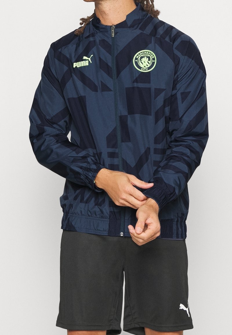Homme ajustant la manche d’une veste Manchester City Puma bleu foncé à motif géométrique, portant un short Puma noir.