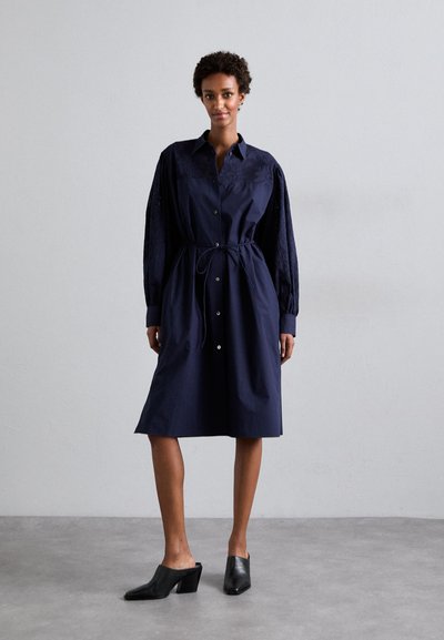 Granatowa sukienka zapinana na guziki z paskiem w talii, z haftowanymi długimi rękawami i spódnicą do kolan. Zestawiona z czarnymi czółenkami na obcasie.