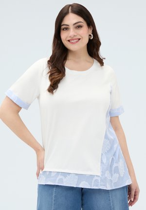 Donna sorridente con capelli scuri indossa una maglietta bianca a maniche corte con dettagli a righe floreali blu e jeans blu, in piedi su sfondo chiaro.