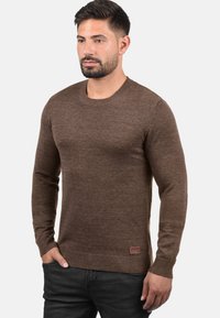 Brun crewneck genser med lange ermer, ribbede mansjetter og fald, myk tekstur og en liten merkelapp nederst til venstre.