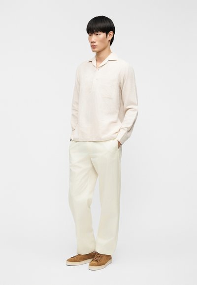 Ανοιχτό μπεζ πουκάμισο από λινό με μακριά μανίκια, γιακά και μία τσέπη στο στήθος, συνδυασμένο με λευκό παντελόνι και καφέ δερμάτινα παπούτσια με κορδόνια.