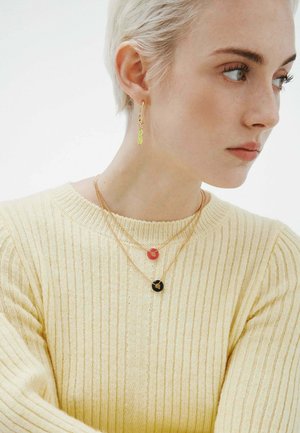 Mujer con cabello corto y rubio, vestida con un suéter acanalado amarillo pálido y collares de oro con colgantes circulares rojos y negros, además de un pendiente de oro.