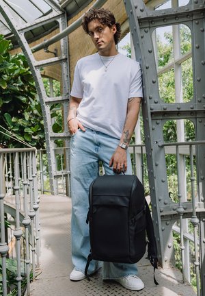 Mann i hvit t-skjorte og jeans som lener seg mot et metallrekkverk, holder en svart ryggsekk i et lyst innendørs hageområde med grøntområder utenfor vinduene.