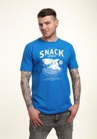 Modré bavlněné tričko s bílým grafickým designem zobrazujícím žraloka a textem "SNACK ATTACK." Krátké rukávy a kulatý výstřih.