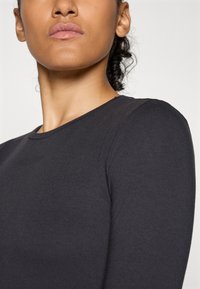 Nahaufnahme einer Person mit mittlerem Hautton, die ein schwarzes Langarmshirt trägt und mit zurückgezogenen lockigen Haaren vor einem schlichten Hintergrund steht.