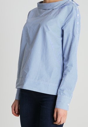 Femme portant un chemisier à manches longues rayé verticalement bleu clair et blanc avec un col montant et un jean bleu foncé.