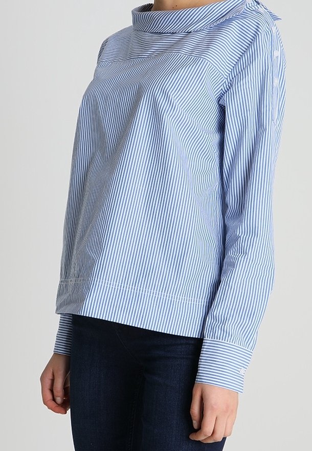 Femme portant un chemisier à manches longues rayé verticalement bleu clair et blanc avec un col montant et un jean bleu foncé.