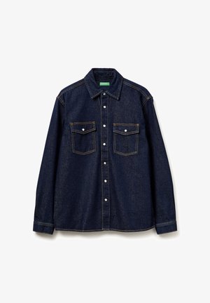 Camisa de mezclilla azul oscura con cuello puntiagudo, dos bolsillos frontales y botones a presión blancos. Presenta costuras visibles y un ajuste relajado.