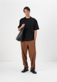 T-shirt noir à manches courtes avec une poche, pantalon ample marron, et chaussures en cuir noires. La personne tient un sac noir.