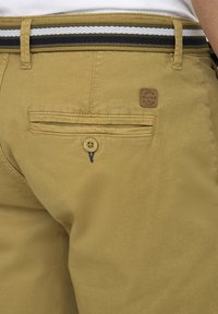 Pantaloni scurți khaki cu o textură netedă, având o curea cu dungi verticale verzi închis și albe, un buzunar pe spate și o etichetă din piele cu marcă.