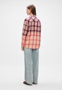 Camisa de xadrez com tons de rosa e bege, mangas longas, detalhe na pala das costas, combinada com calças de ganga largas de tom azul claro e sapatos castanhos com padrão.