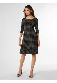 Schwarzes und graues Hahnentrittmuster-Kleid mit Dreiviertelärmeln, einem U-Boot-Ausschnitt und einem ausgestellten Rock, kombiniert mit schwarzen Riemchen-Absatzschuhen.