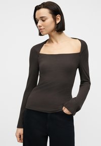 Γυναίκα με κοντά σκούρα μαλλιά, που φορεί στενό, ριγωτό καφέ τοπ με μακριά μανίκια και τετράγωνο ντεκολτέ, καθώς και μαύρο παντελόνι, με το χέρι στην τσέπη, κοιτάζοντας προς τα αριστερά.