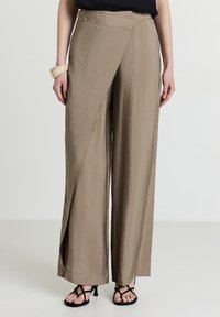 Beige Schlaghose mit strukturierter Oberfläche, versehen mit einem Wickeldesign und einer hohen Taille. Kombiniert mit schwarzen Sandalen mit Riemchen.